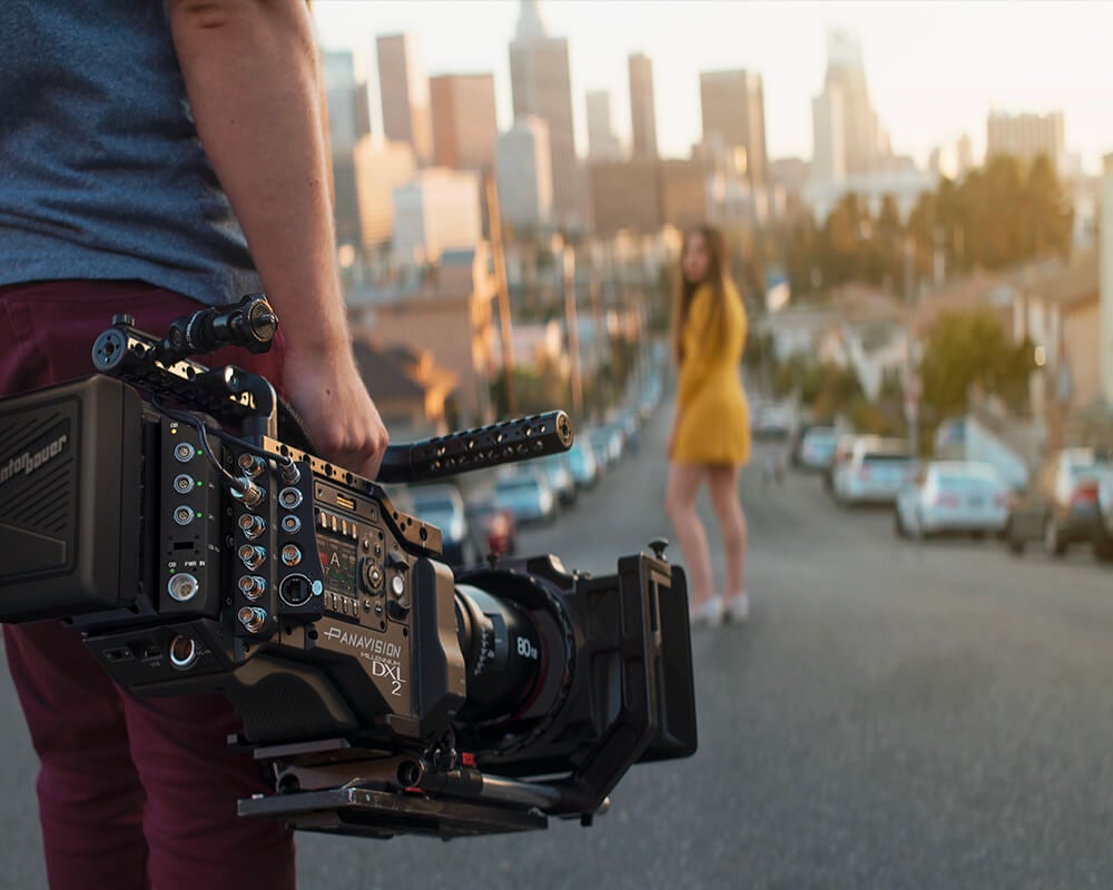 Filmař drží kameru DXL2 namířenou na ženu ve žlutém oblečení stojící na ulici před panoramatem města.